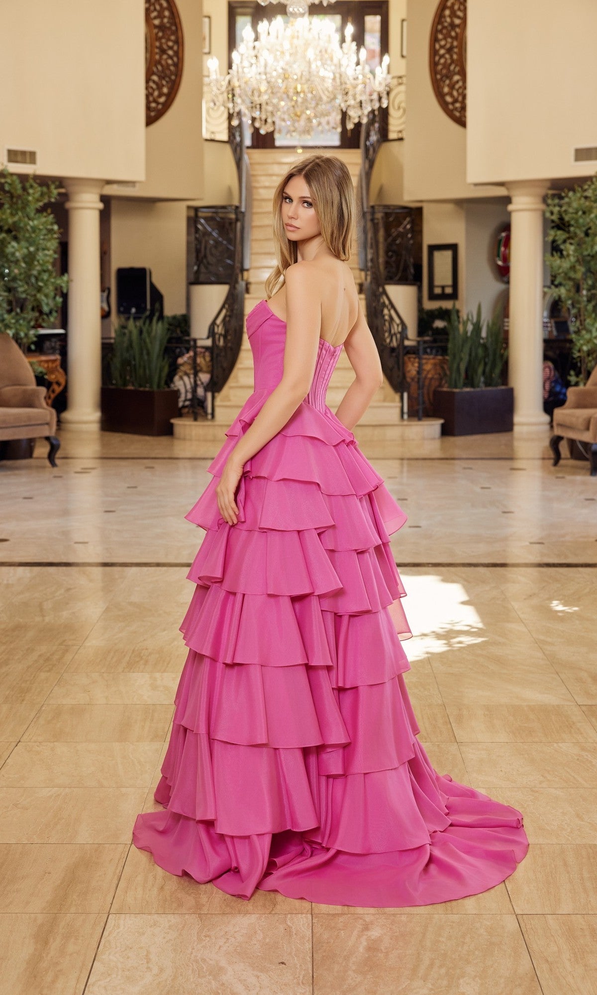 A woman in the Nox Anabel Y1806 long prom dress, featuring a strapless neckline and ruffle tiered skirt, stands indoors on a marble floor with a chandelier and staircase behind her.